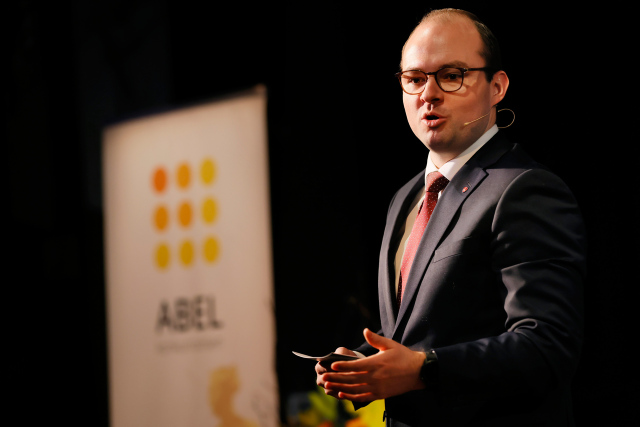 Mann i svart dress som står på scene, med mikrofon (mygg)