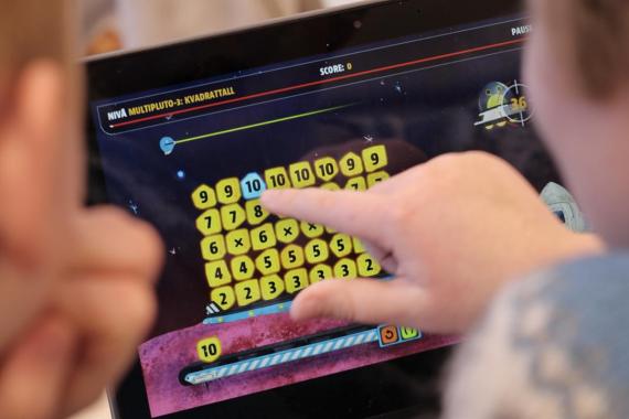 Skjermen til en datamaskin som viser et dataspill med flere tall som ligger oppå hverandre som blokker. I forgrunnen ser vi to barn (blury) og det ene barnet peker på skjermen. 