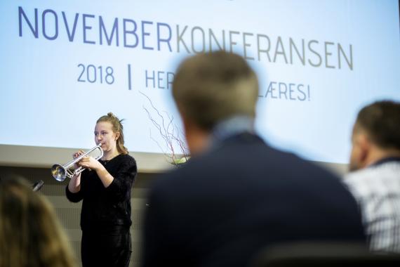Jente som spiller trompet - åpner Novemberkonferansen. I forgrunnen ser vi bakhodet på en mann og bak musikeren, en skjerm med logoen til Novemberkonferansen. 