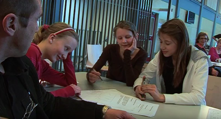 En voksen mann og tre ungdomsskoleelever sitter rundt et bord og diskuterer en oppgave. Det ligger et ark på bordet. 