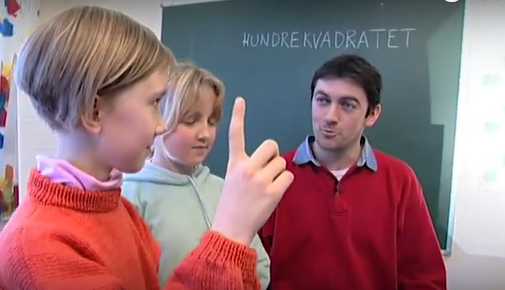 Jente i rød genser som rekker opp hånden. Bak henne sitter en hente i hvit genser, og vi ser lærere i bakgrunnen ved en tavle. På tavla er "hundrekvadratet" skrevet med kritt. 
